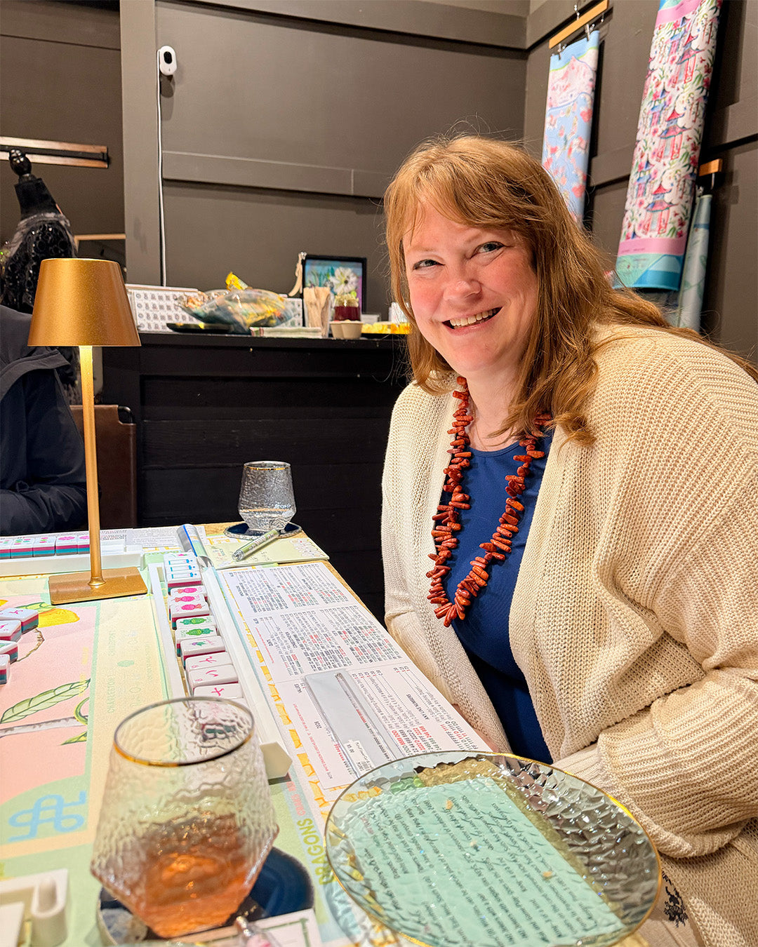 Woman sitting at a mahjong table with a drink smiling.