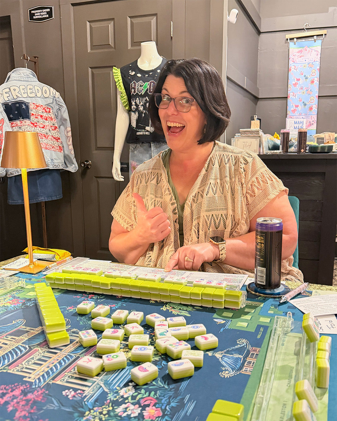 Woman giving a thumbs-up at a mahjong table with snacks and drinks.